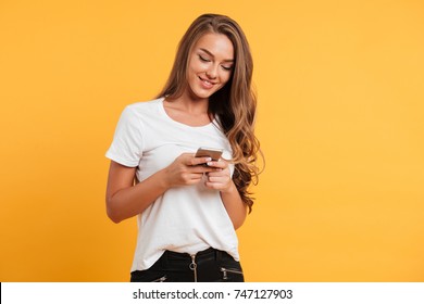 Photo of cheerful cute beautiful young woman chatting by mobile phone isolated over yellow wall background. Looking aside.