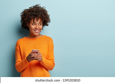 Photo of cheerful delighted African American woman types sms on modern cell phone device, enjoys good internet connection, dressed in orange jumper, focused aside, isolated on blue studio wall