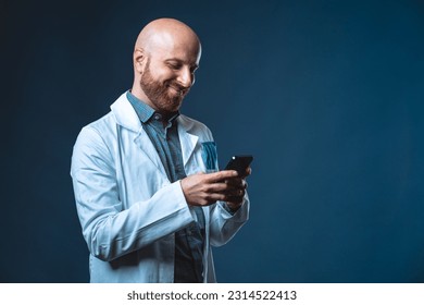 Photo of cheerful doctor with beard smiling and chatting at smart phone with blue background and medical white coat. Medical professional texting sms on cellphone using a social app