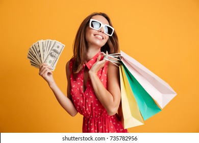 Photo Of Cheerful Elegant Young Woman In Sunglasses And Red Dress Holding Fan Of Money And Colorful Shopping Bags, Looking Aside, Isolated On Yellow Background