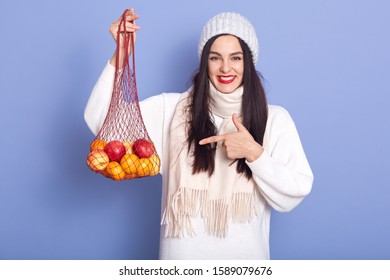 Photo Of Cheerful Good Looking Adorable Young Woman Holding Handbag With Fruit, Directing With Forefinger At It, Standing Isolated Over Lilac Background, Laughing Sincerely. Winter Time Concept.