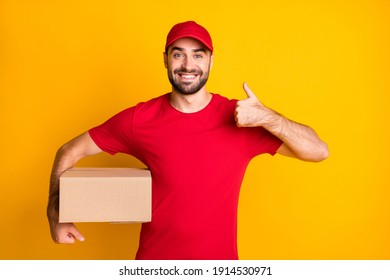 Photo of cheerful guy courier dressed red clothes delivering brown box showing thumb up isolated yellow color background