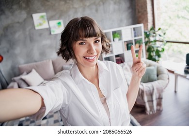 Photo of cheerful happy attractive woman hold hand camera selfie two finger v-sign indoors inside house home