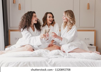 Photo of cheerful and happy three women 20s wearing bathrobe lying in big bed in posh apartment or hotel room and clinking glasses with sparkling wine during bachelorette party