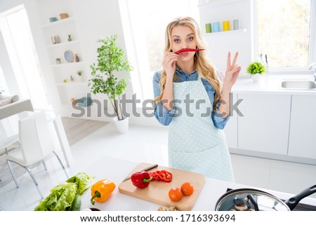 Photo of cheerful housewife blond lady cut salad cooking tasty dinner breakfast hold red hot chilli pepper like mustache showing v-sign symbol playful good mood stand modern kitchen indoors