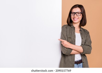 Photo of cheerful lady direct forefinger placard board empty space wear specs khaki shirt isolated beige color background