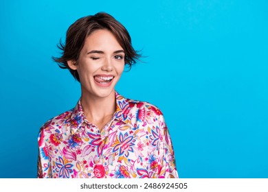 Photo of cheerful lovely girl wear trendy print clothes lick teeth show tongue wink eye empty space isolated on blue color background