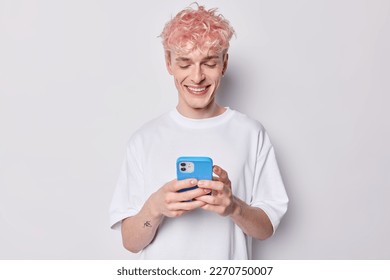 Photo of cheerful man with dyed pink hair laughs at meme his friend sent to him holds mobile phone in hands asnwers on received message dressed in casual t shirt isolated over white background. - Powered by Shutterstock