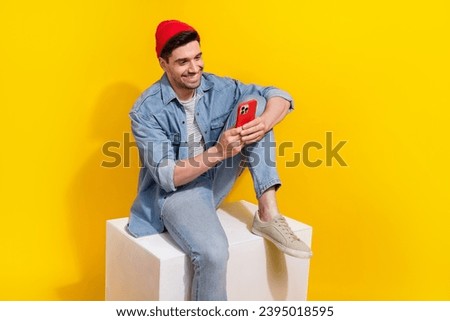 Photo of cheerful positive guy wear denim jacket sitting white platform chatting apple samsung iphone device isolated yellow color background