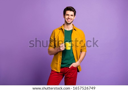 Photo of cheerful positive man with bristle holding hand in pocket of trousers smiling toothily holding cup of coffee isolated violet pastel color background
