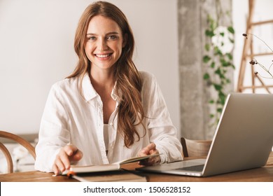 Photo of a cheerful positive young pretty business woman sit indoors in office using laptop computer holding notebook.
