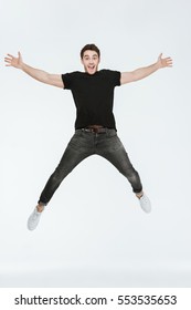 Photo of cheerful young man dressed in black t-shirt jumping over white background looking at camera.
