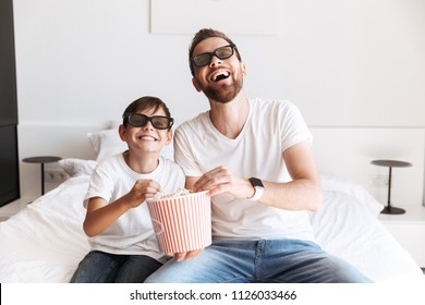 Photo of cheerful young man father dad having fun with his son indoors at home watching TV eating pop corn wearing 3D glasses.