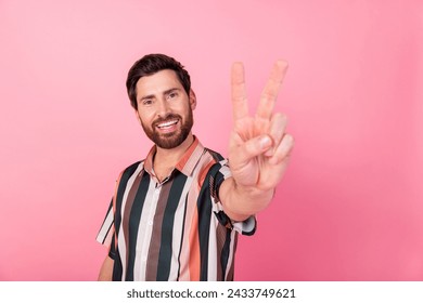 Photo of cheerful young man toothy smile hand fingers demonstrate v-sign isolated on pink color background