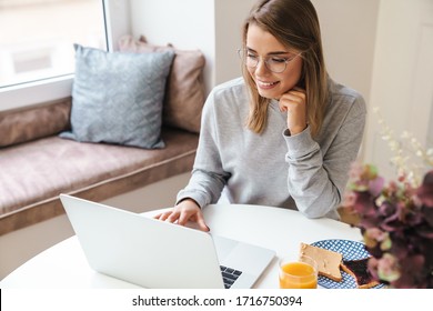 Photo of cheerful young woman in eyeglasses using laptop while having breakfast at living room - Powered by Shutterstock