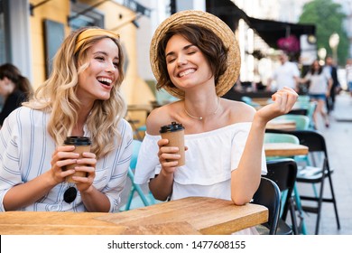 Photo of a cheery laughing young girls friends sitting outdoors in cafe drinking coffee talking with each other.