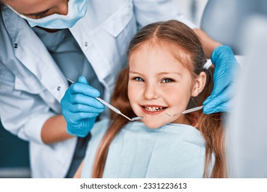 Photo of a child smiling at a dentist appointment. A doctor in gloves holds examination tools behind. Children's dentistry. - Powered by Shutterstock