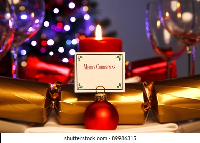 Photo Of A Christmas Dinner Place Setting And Bauble Card Holder With Decorated Tree Out Of Focus In The Background. Lit By Candlelight.