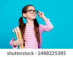 Photo of clever small girl with ponytails dressed striped shirt in glasses holding book look empty space isolated on blue color background