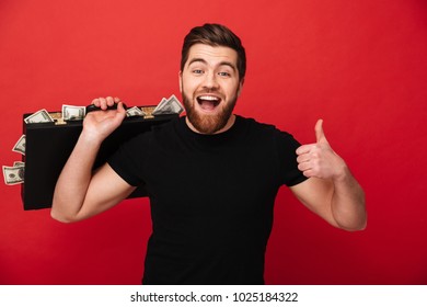 Photo close up of positive bearded man in black t-shirt expressing joy while carrying briefcase full of dollar bills isolated over red wall