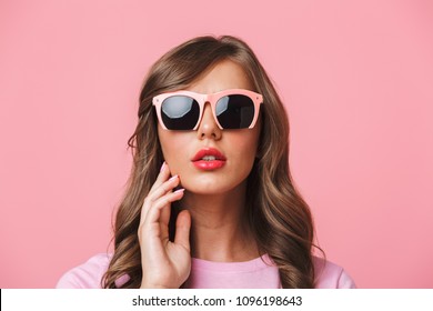 Photo closeup of sexual charming woman 20s with long curly hairstyle wearing trendy sunglasses looking at camera with fashion look isolated over pink background