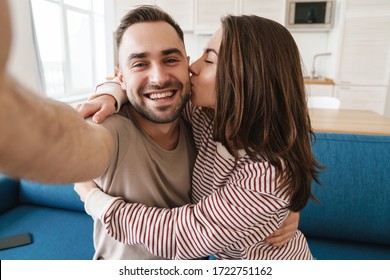 Photo closeup of young joyful couple hugging and kissing while taking selfie photo in kitchen - Powered by Shutterstock