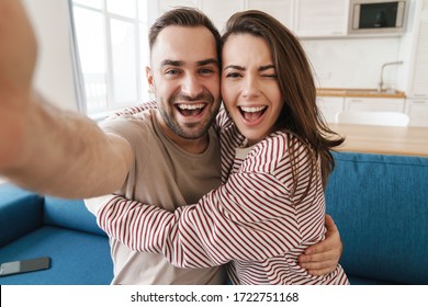 Photo closeup of young joyful couple hugging and winking while taking selfie photo in kitchen - Powered by Shutterstock