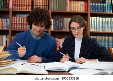 Photo The College Students' Learning Process. A Young Guy And A Girl Sit In The Library Near Books And Notebooks, Doing Work On The Project. High Quality Photo