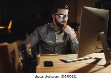 Photo Of Concentrated Bearded Web Designer Dressed In Shirt Working Late At Night And Looking At Computer.