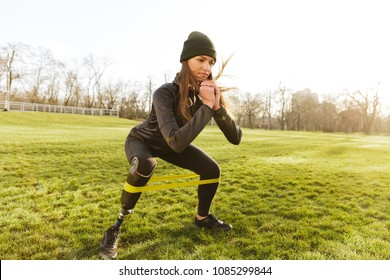 Photo of concentrated disabled girl in tracksuit doing sports and squatting with prosthetic leg on grass using resistance band - Powered by Shutterstock