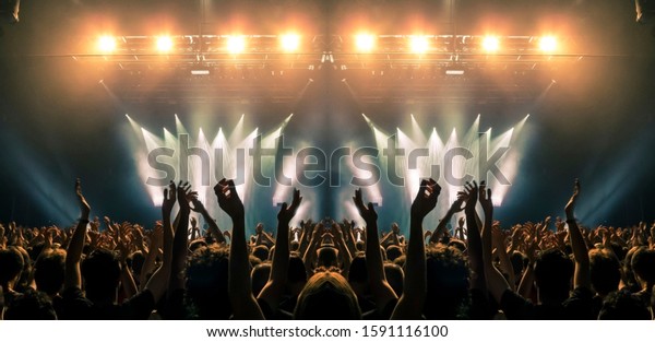 Photo of a concert hall\
with people silhouettes clapping in front of a big stage lit by\
spotlights. Shot is taken from concert crowd point of view, lens\
flare is visible.