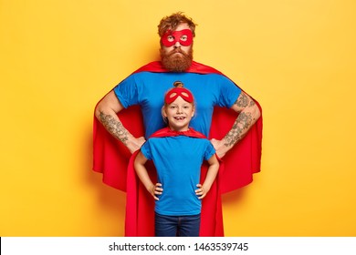 Photo of confident ginger father and daughter keep hands on waists, pretend being superhero and defend people, wear special costumes, isolated on yellow background. Family team ready to work