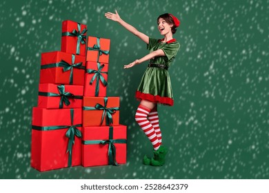 Photo of confident impressed young woman dressed elf costume showing arms gift boxes pile smiling isolated green color background - Powered by Shutterstock