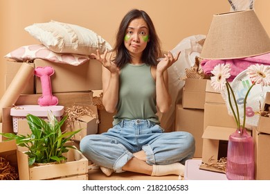 Photo of confused young Asian woman shrugs shoulders sits crossed legs feels unaware applies green hydrogel patches under eyes to reduce wrinkles relocates to new apartment. Removal concept.