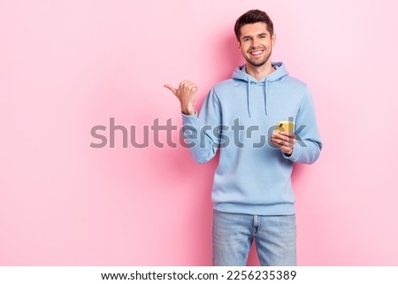 Photo of cool sweet man wear blue hoodie writing sms modern device pointing thumb empty space isolated pink color background