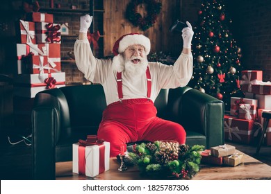Photo of crazy addicted grey beard santa claus sit ouch have funny x-mas noel party play video game win raise fists wear red cap headwear in house indoors with christmas evergreen tree decoration
