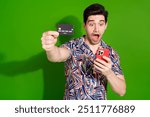 Photo of crazy guy with stylish haircut dressed print shirt staring at smartphone show you credit card isolated on green color background