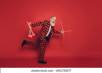 Photo Of Crazy Retired Cupid Hold Arrows Bow Run Open Mouth Wear Heart Print Tux Tie Shoes Isolated Red Background