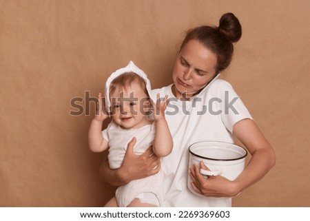 Photo of crying mother wearing white T- shirt holding her toddler daughter in hands and pot, cooking with infant baby, talking phone and being stressed, posing isolated over brown background.