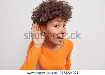 Photo of curious young woman keeps hand near ear bends forward eavesdropes tries to overhear interesting information cant hear you listens someones conversation wears orange sweater poses indoor
