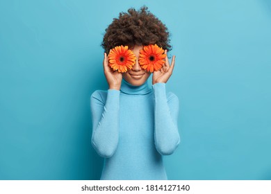 Photo of curly haired female florist has fun while makes decorations for special occasion, keeps two orange gerbera daisies in front of eyes and wears casual turtleneck. Women flowers concept - Shutterstock ID 1814127140