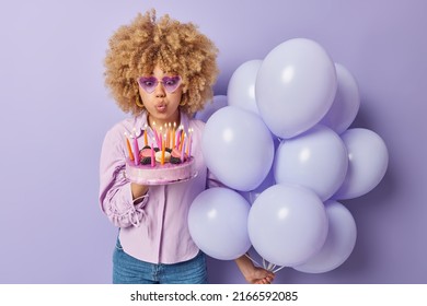 Photo of curly haired woman blows candles on cake holds bunch of inflated balloons wears sunglasses and shirt isolated over purple background. People celebration and festive occasion concept