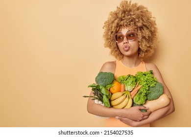 Photo Of Curly Haired Woman Carries Bunch Of Fresh Fruit And Vegetables Keeps Lips Folded Wears Trendy Sunglasses And Casual T Shirt Isolated Over Brown Background Blank Copy Space For Promotion