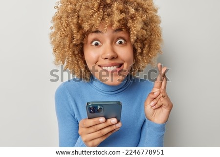 Photo of curly woman holds mobile phone crosses fingers makes wish anticipates something hopes to receive something dressed in casual blue turtleneck isolated on grey background believes in good luck
