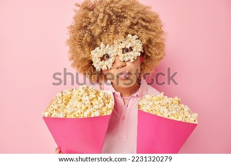 Photo of curly woman wears handmade creative eyeglasses poses with delicious popcorn attends cinema together with friend keeps lips rounded watches interesting movie isolated over pink wall.