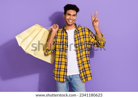 Photo of cute cheerful guy dressed checkered yellow shirt showing v-sign holding bargains isolated purple color background