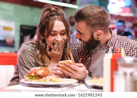 Photo of a cute happy optimistic young loving couple in retro bright street food cafe using mobile phone.