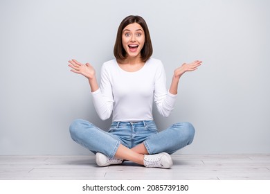 Photo of cute impressed young woman wear white outfit rising arms sitting legs crossed smiling isolated concrete grey wall background