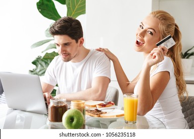 Photo of cute smiling young loving couple have a breakfast indoors at home while using laptop computer holding credit card. - Shutterstock ID 1056235118