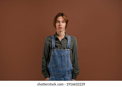 Photo Of Cute Woman Farmer Stands With Serious Face. Female Wears Denim Overalls, Isolated Brown Color Background